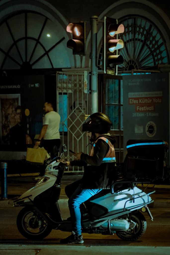 Motorcyclist on a scooter at night in an urban setting with traffic lights and pedestrians.